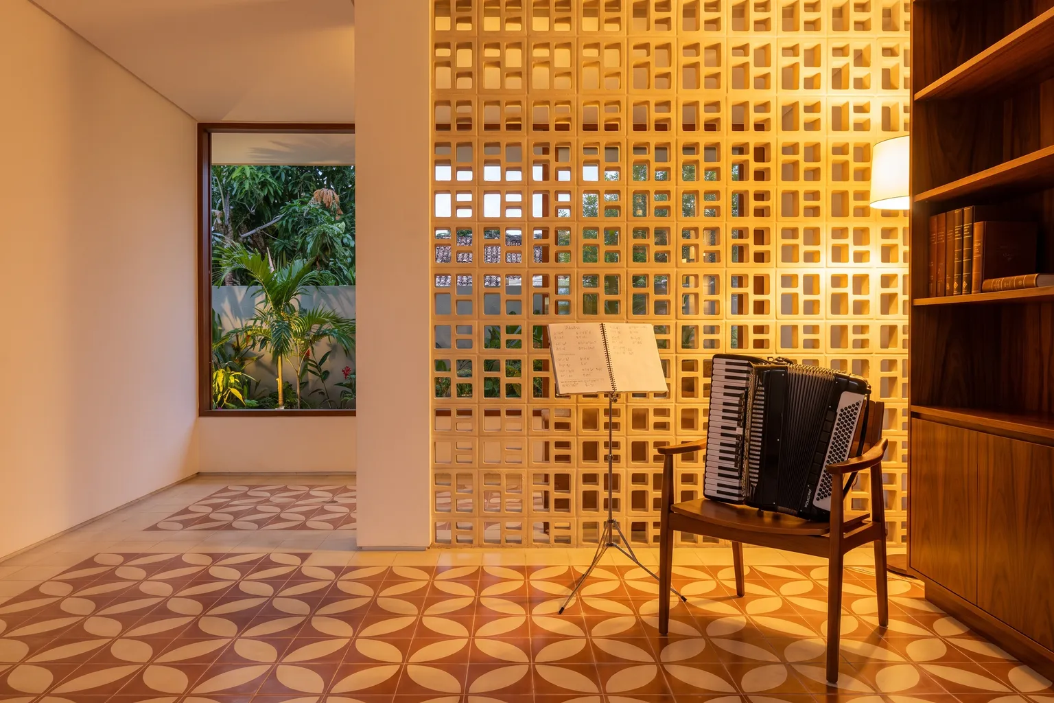 Sala de música com parede de cobogó iluminada, estante de madeira, uma estante de partitura e uma sanfona numa cadeira